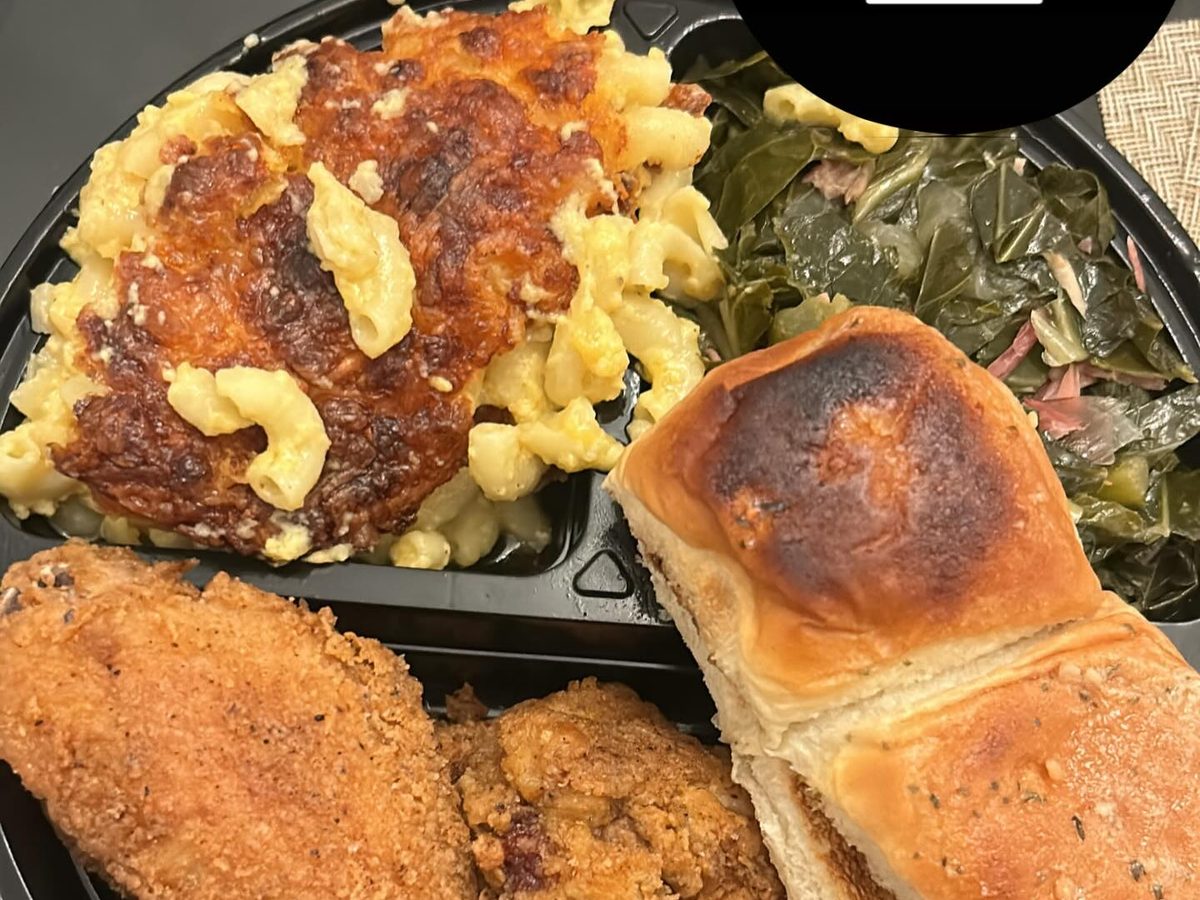 Fried chicken, baked mac and cheese with a browned crust, collard greens and a toasted garlic butter roll in a black takeout tray